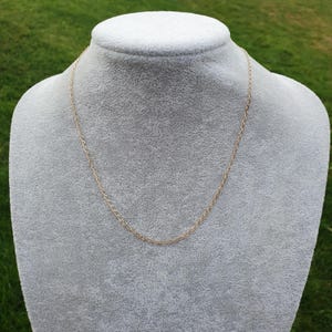 May include: A delicate gold-colored chain necklace displayed on a gray mannequin. The chain is made of small, interconnected links, creating a simple yet elegant design. The necklace is a classic piece of jewelry.