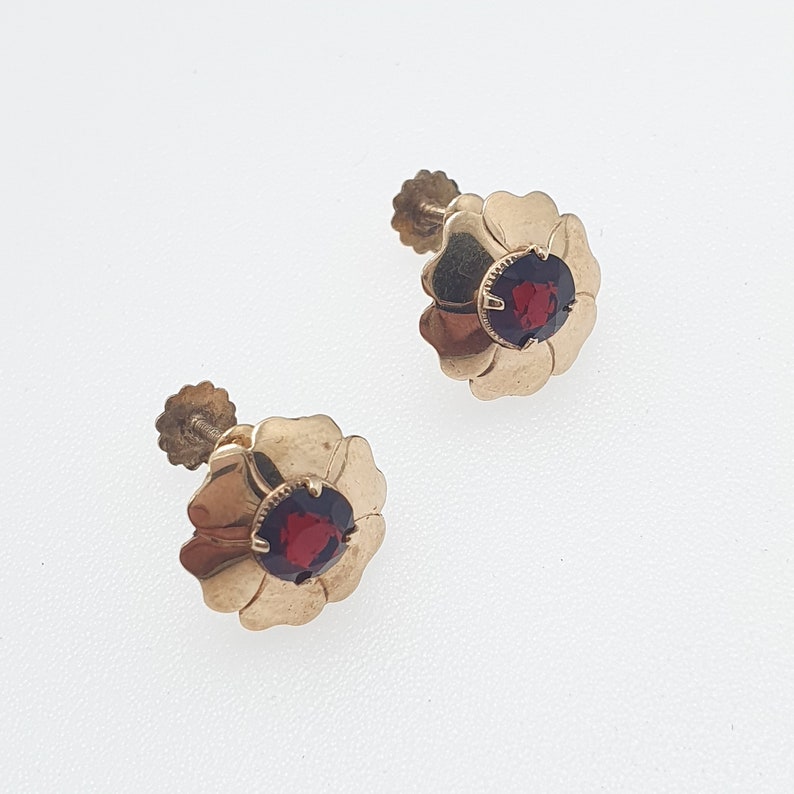 May include: A pair of gold earrings with red gemstone accents. Each earring features a flower-shaped design with a round red gemstone in the centre.