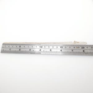 May include: A delicate gold-colored chain necklace with a small clasp, laid out on a ruler. The chain is approximately 18 inches long. The necklace is on a white background, showcasing its simple elegance and design.