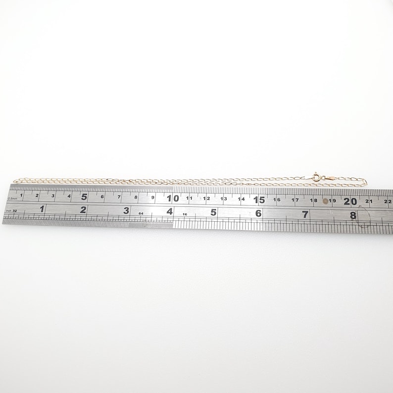 May include: A delicate gold-colored chain necklace with a small clasp, laid out on a ruler. The chain is approximately 18 inches long. The necklace is on a white background, showcasing its simple elegance and design.