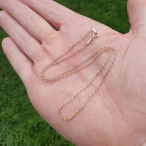 May include: A delicate gold-colored chain necklace with an oval link design. The necklace is displayed on a hand against a backdrop of green grass. The clasp is visible at the top of the necklace.