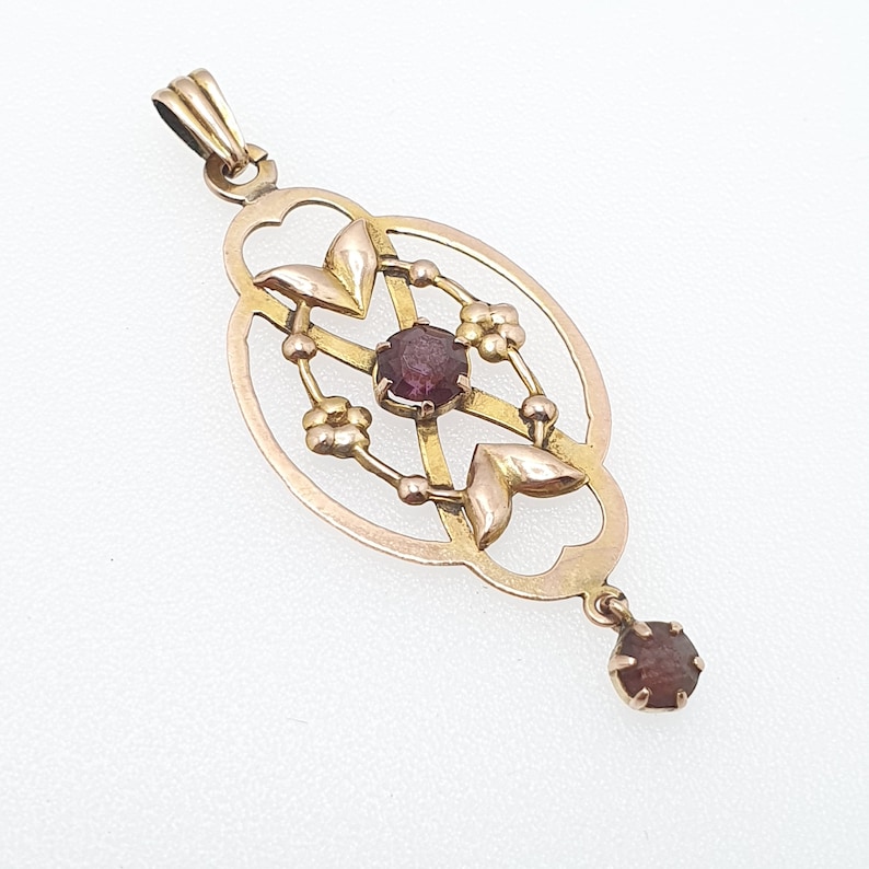 Pode incluir: Um pingente de ouro com um design delicado que apresenta uma pedra preciosa central roxa e pequenos detalhes florais. O pingente est&aacute; suspenso em uma corrente de ouro.