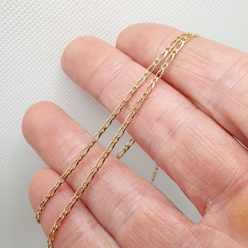 May include: A gold chain necklace with a delicate, figaro style chain. The chain is shown laying on a hand.