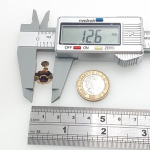 May include: A close-up of a gold flower-shaped earring with a red gemstone in the centre. The earring is being measured with a caliper, which reads 12.6 mm. A British one pound coin is also in the image for scale.