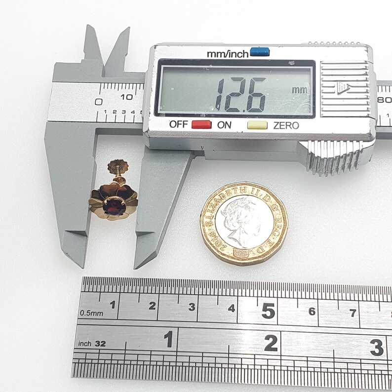 May include: A close-up of a gold flower-shaped earring with a red gemstone in the centre. The earring is being measured with a caliper, which reads 12.6 mm. A British one pound coin is also in the image for scale.
