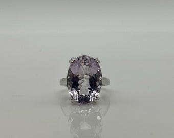 May include: A silver ring with a large, oval-shaped, light purple gemstone. The gemstone is held in place by four prongs. The ring's band is polished silver. The ring is shown on a reflective surface, creating a mirrored effect.