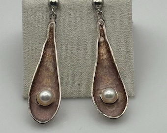 May include: A pair of silver-toned drop earrings. Each earring features a teardrop-shaped design with a pearl at the base. The interior has a bronze hue, and the outer edge is a light pink color. The earrings are displayed on a gray surface.