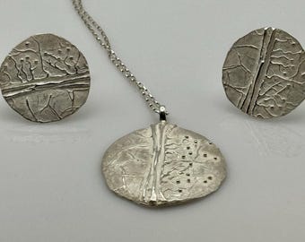 Puede incluir: Un conjunto de joyas de plata con un collar con colgante y pendientes a juego. El colgante y los pendientes tienen un diseño texturizado y orgánico con un patrón similar a un árbol. La cadena del collar es delicada.