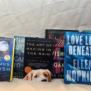 K&ouml;nnte beinhalten: Eine Sammlung von Hardcover-B&uuml;chern mit verschiedenen Titeln und Autoren. Die Buchumschl&auml;ge zeigen eine Reihe von Farben, darunter Blau, Schwarz und Gr&uuml;n. Ein Hund schaut hinter den B&uuml;chern hervor. Die B&uuml;cher sind auf einer wei&szlig;en Oberfl&auml;che angeordnet.