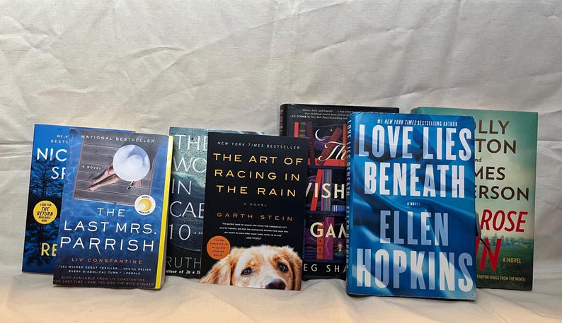 K&ouml;nnte beinhalten: Eine Sammlung von Hardcover-B&uuml;chern mit verschiedenen Titeln und Autoren. Die Buchumschl&auml;ge zeigen eine Reihe von Farben, darunter Blau, Schwarz und Gr&uuml;n. Ein Hund schaut hinter den B&uuml;chern hervor. Die B&uuml;cher sind auf einer wei&szlig;en Oberfl&auml;che angeordnet.