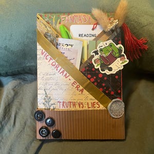 K&ouml;nnte beinhalten: Eine dekorative Box mit den W&ouml;rtern "FANTASY" und "VICTORIAN-ERA". Die Box hat ein goldenes Band, einen Froschaufkleber und mehrere Kn&ouml;pfe. Sie enth&auml;lt auch einen Stift, ein Teebeutel und ein Etikett mit der Aufschrift "READING".