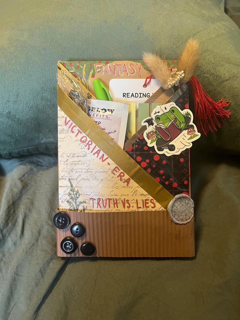 K&ouml;nnte beinhalten: Eine dekorative Box mit den W&ouml;rtern "FANTASY" und "VICTORIAN-ERA". Die Box hat ein goldenes Band, einen Froschaufkleber und mehrere Kn&ouml;pfe. Sie enth&auml;lt auch einen Stift, ein Teebeutel und ein Etikett mit der Aufschrift "READING".