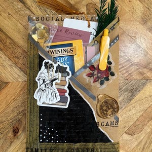 K&ouml;nnte beinhalten: Ein handgefertigtes Journal mit einem Collage-Design. Das Journal enth&auml;lt eine goldene Quaste, einen Aufkleber einer lesenden Frau und einen Teebeutel. Die W&ouml;rter "SOCIAL MEDIA" und "BOOK REVIEW" sind sichtbar.