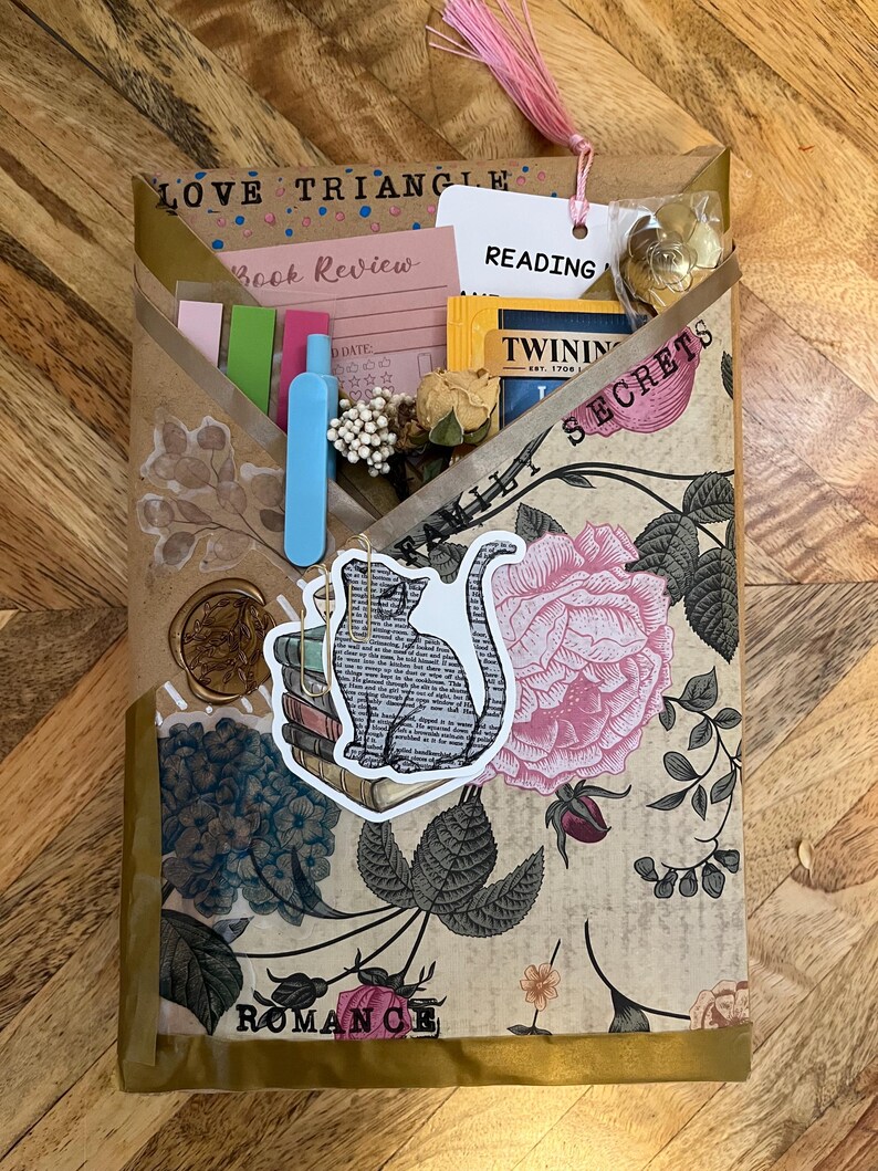 K&ouml;nnte beinhalten: Eine handgefertigte Bucheinschlagh&uuml;lle mit floralem und botanischem Design, mit einem Katzenaufkleber auf B&uuml;chern. Die H&uuml;lle enth&auml;lt einen rosa Quastenanh&auml;nger, Etiketten und kleine Gegenst&auml;nde. Die W&ouml;rter "Love Triangle", "Book Review", "Reading", "Secrets" und "Romance" sind sichtbar.