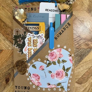 K&ouml;nnte beinhalten: Eine handgefertigte Bucheinschlagh&uuml;lle mit floralem Herzdesign, mit den Worten "YOUNG ADULT" und "ROMANCE". Enth&auml;lt einen Lesezeichen, Aufkleber, Tee und einen blauen Stift. Die H&uuml;lle besteht aus braunem Papier und ist mit einer goldenen Blume und einem Wachssiegel verziert.