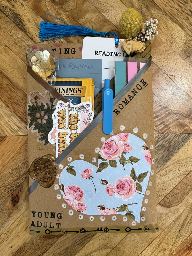 K&ouml;nnte beinhalten: Eine handgefertigte Bucheinschlagh&uuml;lle mit floralem Herzdesign, mit den Worten "YOUNG ADULT" und "ROMANCE". Enth&auml;lt einen Lesezeichen, Aufkleber, Tee und einen blauen Stift. Die H&uuml;lle besteht aus braunem Papier und ist mit einer goldenen Blume und einem Wachssiegel verziert.