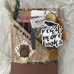 K&ouml;nnte beinhalten: Eine braune Papiertasche mit den Aufschriften "WORLD WAR II" und "HISTORICAL FICTION". Die Tasche enth&auml;lt einen Stift, Lesezeichen, einen Teebeutel und einen Aufkleber mit der Aufschrift "read more books". Dekorative Elemente sind getrocknete Pflanzen und eine goldfarbene Quaste.