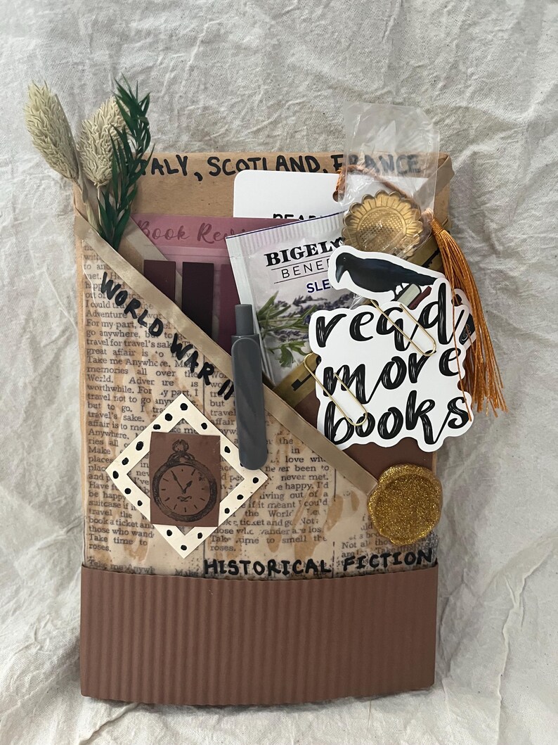 K&ouml;nnte beinhalten: Eine braune Papiertasche mit den Aufschriften "WORLD WAR II" und "HISTORICAL FICTION". Die Tasche enth&auml;lt einen Stift, Lesezeichen, einen Teebeutel und einen Aufkleber mit der Aufschrift "read more books". Dekorative Elemente sind getrocknete Pflanzen und eine goldfarbene Quaste.