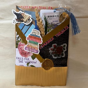 K&ouml;nnte beinhalten: Eine handgefertigte Geschenkbox mit goldfarbener Wellpappe und schwarzem Deckel. Die Box ist mit Aufklebern verziert, darunter ein Stapel B&uuml;cher, eine Rose und ein Rabe. Der Text auf der Box lautet "FLEES APUSIVE EX", "ROMANCE THRILLER" und "FALLS FOR THE WIDOW".