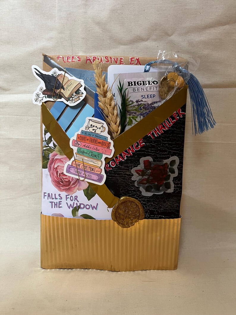K&ouml;nnte beinhalten: Eine handgefertigte Geschenkbox mit goldfarbener Wellpappe und schwarzem Deckel. Die Box ist mit Aufklebern verziert, darunter ein Stapel B&uuml;cher, eine Rose und ein Rabe. Der Text auf der Box lautet "FLEES APUSIVE EX", "ROMANCE THRILLER" und "FALLS FOR THE WIDOW".