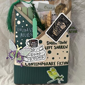K&ouml;nnte beinhalten: Eine dekorative Buchbox mit dem Text "NAVIGATE GRIEF" und "UNLIKELY ALLIES". Sie zeigt Aufkleber eines Schmetterlings, einer Libelle und eines B&uuml;cherstapels mit den Worten "GOOD BOOKS" und "CONTEMPORARY FICTION".