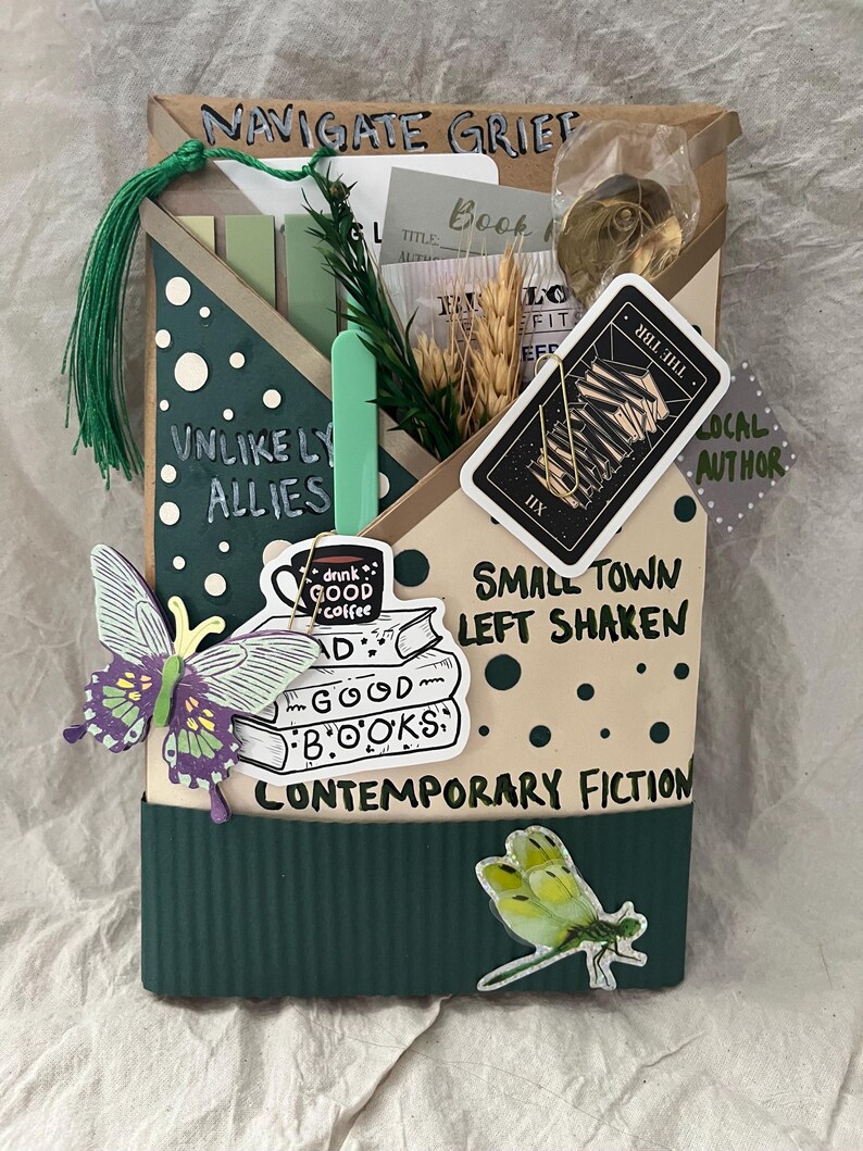 K&ouml;nnte beinhalten: Eine dekorative Buchbox mit dem Text "NAVIGATE GRIEF" und "UNLIKELY ALLIES". Sie zeigt Aufkleber eines Schmetterlings, einer Libelle und eines B&uuml;cherstapels mit den Worten "GOOD BOOKS" und "CONTEMPORARY FICTION".