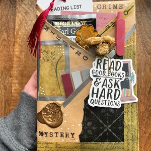 K&ouml;nnte beinhalten: Dekorative Geschenkverpackung mit den W&ouml;rtern "Mystery" und "Crime". Enth&auml;lt einen Teebeutel, einen Lesezeichen mit "Reading List" und einen Aufkleber mit "Read Good Books & Ask Hard Questions". Mit roter Quaste.