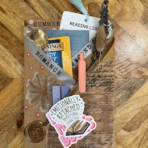 K&ouml;nnte beinhalten: Ein handgemachtes Geschenkpaket mit Lese-Thema. Es enth&auml;lt einen Teebeutel, ein Lesezeichen, einen Aufkleber mit der Aufschrift "Emotionally Attached to Fictional Characters" und einen kleinen orangefarbenen Stift. Das Paket ist mit einem Band mit der Aufschrift "Romance" verziert.