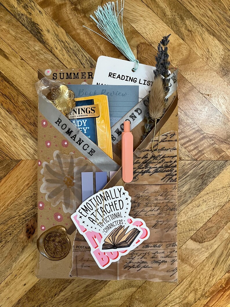 K&ouml;nnte beinhalten: Ein handgemachtes Geschenkpaket mit Lese-Thema. Es enth&auml;lt einen Teebeutel, ein Lesezeichen, einen Aufkleber mit der Aufschrift "Emotionally Attached to Fictional Characters" und einen kleinen orangefarbenen Stift. Das Paket ist mit einem Band mit der Aufschrift "Romance" verziert.
