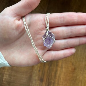 May include: A handcrafted necklace featuring a raw, purple amethyst crystal pendant. The crystal is encased in a woven, off-white macrame cord, suspended from a matching cord necklace. The natural stone has a rough texture and varying shades of purple.