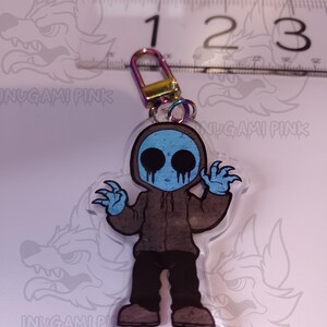 May include: A clear acrylic keychain featuring a cartoon character with a blue face, brown hoodie, and black pants. The character has a creepy smile and is holding out its hands. The keychain has a metal ring attached to the top.