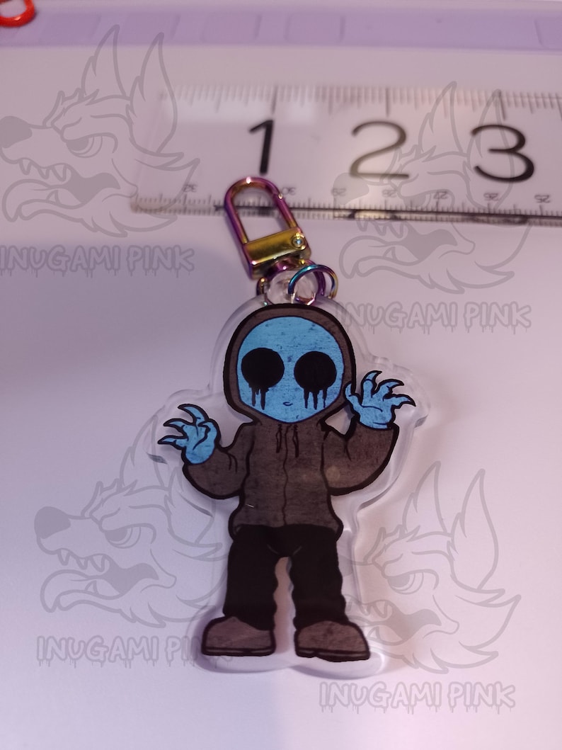 May include: A clear acrylic keychain featuring a cartoon character with a blue face, brown hoodie, and black pants. The character has a creepy smile and is holding out its hands. The keychain has a metal ring attached to the top.