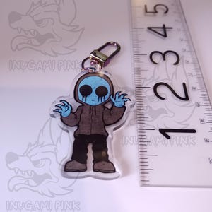 May include: A blue and brown cartoon character with a creepy smile and a blue hooded sweatshirt. The character is holding a key chain with a gold metal ring. The character is approximately 2 inches tall.
