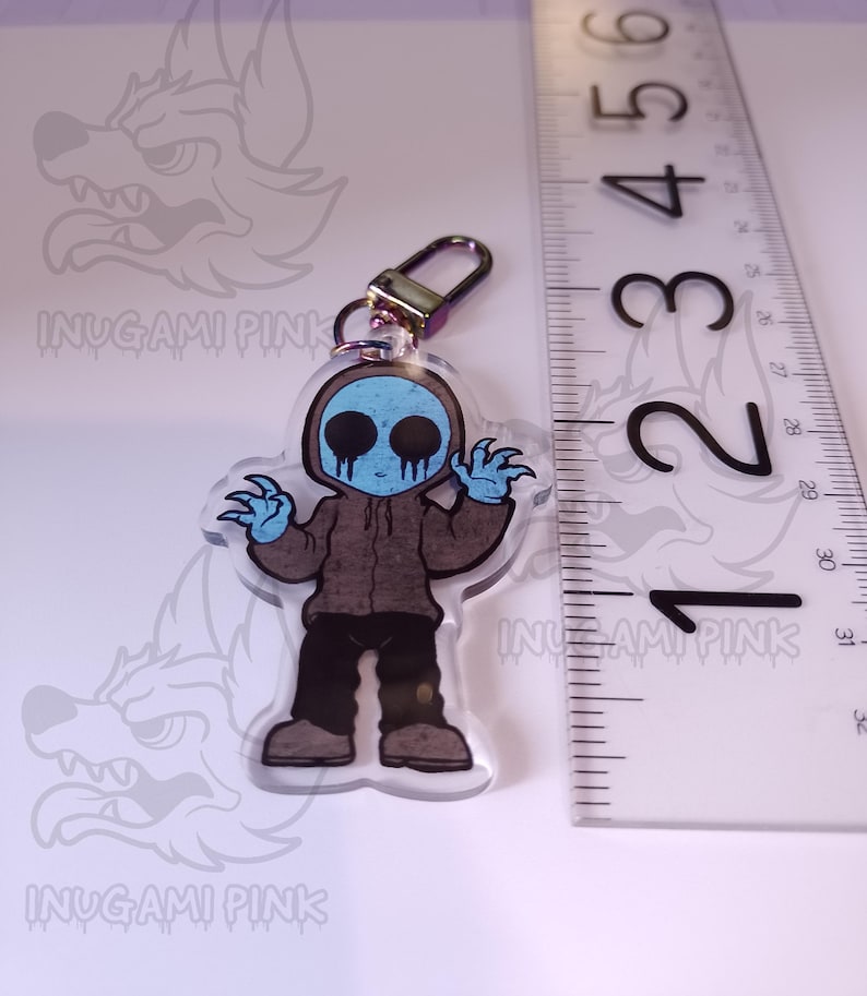 May include: A blue and brown cartoon character with a creepy smile and a blue hooded sweatshirt. The character is holding a key chain with a gold metal ring. The character is approximately 2 inches tall.