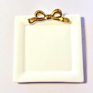May include: A square, off-white ceramic plate with a gold-colored bow accent at the top. The plate has a slightly raised edge and a smooth surface. The bow is detailed with curved lines and a textured appearance, adding a touch of elegance.
