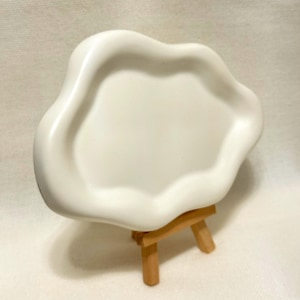 May include: A white, organically shaped ceramic tray with a wavy rim. The tray is displayed on a small, natural wood easel. The design is minimalist and modern, suitable for decorative or functional use.