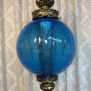 May include: A vintage hanging light fixture featuring a large, textured, blue glass globe. The globe is encased in ornate, gold-toned metalwork at the top and bottom, with decorative accents. The fixture hangs from a chain.