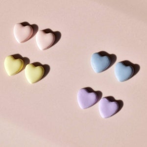 May include: A collection of pastel-colored heart-shaped candies arranged on a light pink surface. The candies are in pairs of pink, yellow, blue, and purple. The image is well-lit, with soft shadows.