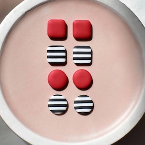 Może przedstawiać: Kolekcja ręcznie robionych kolczyków w różnych kształtach i wzorach. Kolczyki obejmują pary czerwonych kwadratów, czarno-białych kwadratów w paski, czerwonych kół i czarno-białych kół w paski. Kolczyki są prezentowane na różowym talerzu.