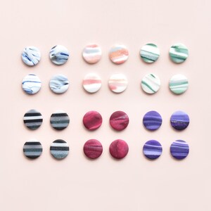 May include: A collection of colorful, round stud earrings arranged on a light pink surface. The earrings feature various designs, including marbled patterns in blue, pink, and green, as well as striped and solid color options.