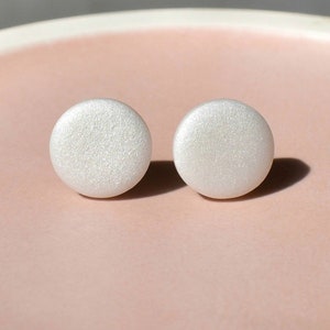 Könnte beinhalten: Ein Paar runde, weiße Ohrstecker mit strukturiertem, perlmuttartigem Finish. Die Ohrringe werden auf einer hellrosa Oberfläche präsentiert, wodurch eine weiche, feminine Ästhetik entsteht. Ein schlichtes, aber elegantes Accessoire.