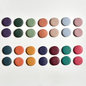 Op de afbeelding: Een verzameling van vierentwintig ronde houten knopen in verschillende kleuren, waaronder paars, groen, oranje en roze. De knopen zijn in een rasterpatroon op een witte achtergrond gerangschikt, met een divers palet aan tinten.