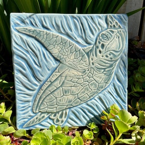 May include: A ceramic tile with a blue and white glaze depicting a sea turtle swimming in the ocean. The turtle is in relief and has a detailed shell and flippers.