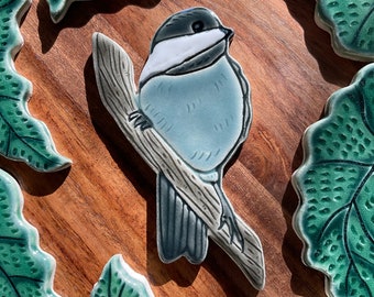May include: A blue and white ceramic bird perched on a branch, surrounded by green leaves. The bird has a black eye and a white chest. The leaves are detailed with a textured pattern.
