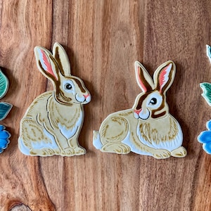 May include: Two ceramic rabbit figurines with brown and white fur, painted with details of brown, white, and green. The rabbits are sitting on a wooden surface.