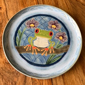 May include: A round ceramic plate featuring a central embossed design of a green frog with red eyes perched on a brown branch. The background shows a blue grid pattern with three purple flowers. The plate has a light blue and white border.