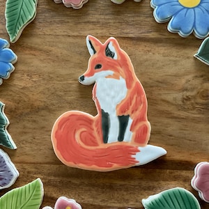 May include: A ceramic fox figurine with orange, white, and black details. The fox is surrounded by floral and leaf-shaped ceramic pieces in various colors, including blue, pink, and green. The items are arranged on a wooden surface.
