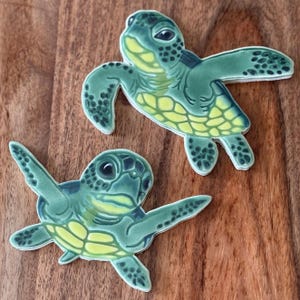 May include: Two ceramic sea turtle figurines. The turtles are primarily green with yellow underbellies and black detailing. The turtles are on a wooden surface.