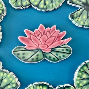 May include: A pink water lily with green leaves, painted on a ceramic tile. The tile is a light blue color.