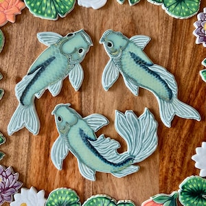 May include: Three teal koi fish cookies with detailed fins and dark blue accents are arranged on a wooden surface. The cookies are surrounded by decorative floral and lily pad cookies. The koi fish have a watercolor effect.